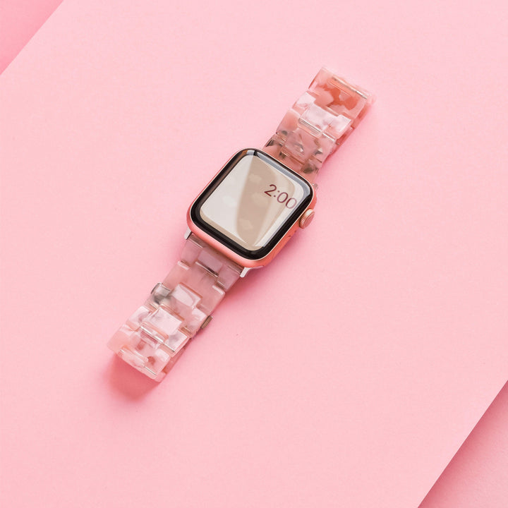 A girly and cute smartwatch with a rectangular face and rose gold casing is displayed on a pink background. The Luxe Pink Clouds Apple Watch Strap by Coconut Lane features a pink and white marbled pattern made of durable resin. The screen shows the time as 2:00.