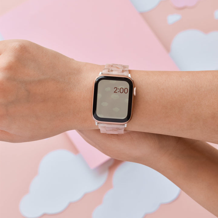 A person wearing the Luxe Pink Clouds Apple Watch Strap by Coconut Lane shows the time as 2:00. The background consists of pastel pink and white cloud-like shapes. Only the wrist and hand of the person are visible, with the smartwatch prominently displayed.