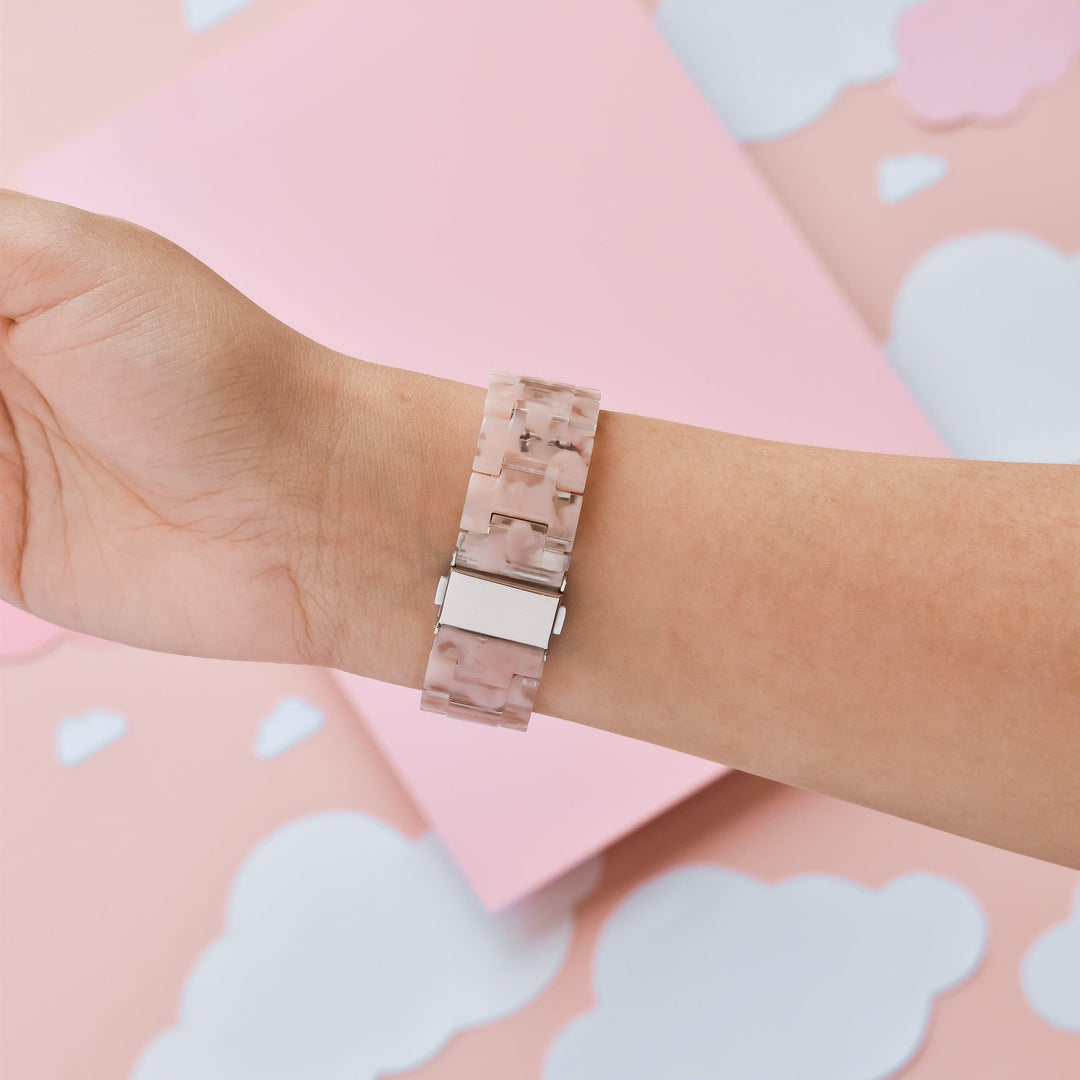 A hand is shown from the wrist up, wearing the Luxe Pink Clouds Apple Watch Strap by Coconut Lane, which features a pink marbled design and a durable resin rectangular silver clasp. The background presents soft pastel pink and white paper clouds for a girly and cute aesthetic.