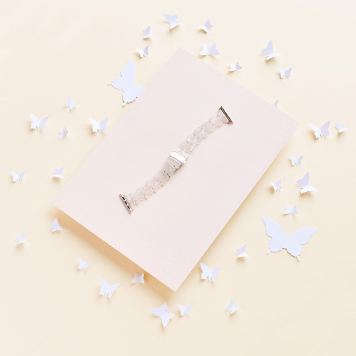 The Luxe Holo Butterfly Apple Watch Strap by Coconut Lane is showcased on a light beige rectangular board, surrounded by scattered holographic butterflies of various sizes. The white beaded strap creates a delicate and airy composition against the soft, pale yellow background.