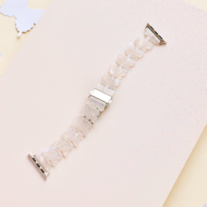 A watch band made of small, translucent, white, butterfly-shaped links with silver buckle clasps is laid out on a lightly textured, pale pink surface. A white butterfly cutout is partially visible in the top left corner. This adjustable Luxe Holo Butterfly Apple Watch Strap by Coconut Lane features delicate holographic butterflies for a whimsical touch.