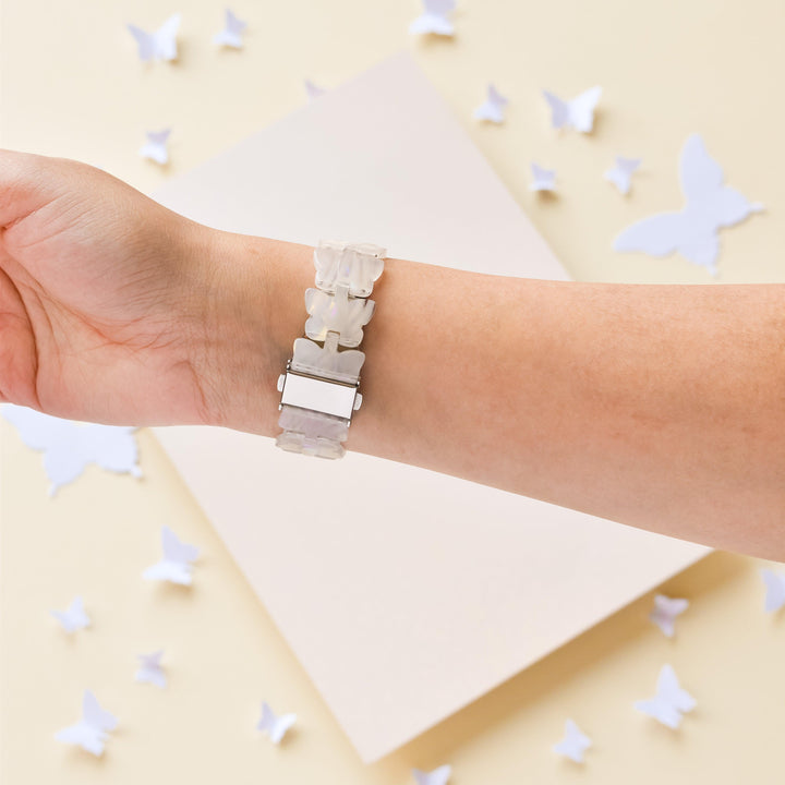 A hand is shown wearing the Luxe Holo Butterfly Apple Watch Strap by Coconut Lane, featuring a light-colored, decorative design. The background showcases cut-out white butterfly shapes scattered on a neutral surface, creating a delicate and whimsical atmosphere.