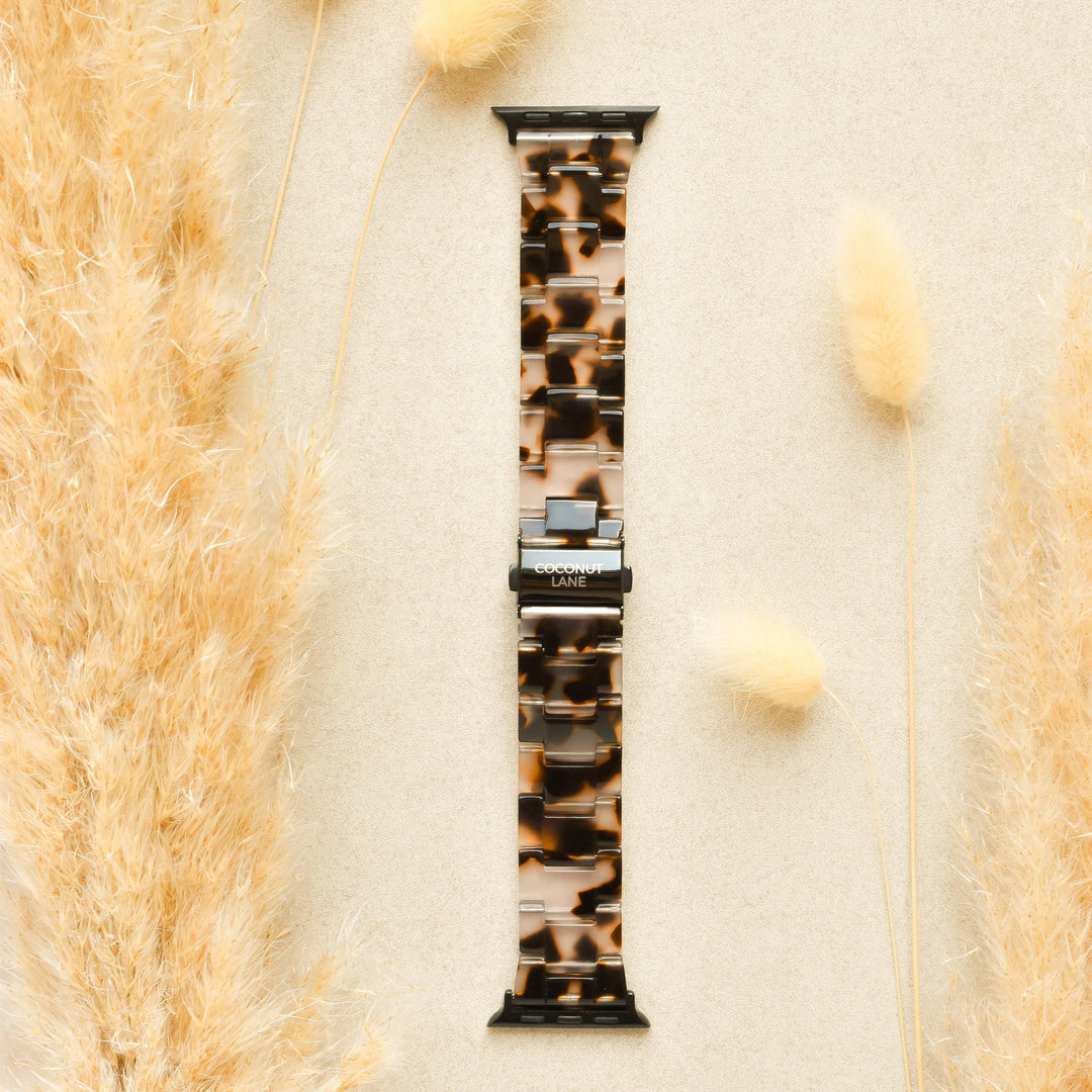 A Luxe Ivory Tort Apple Watch Strap from Coconut Lane is placed on a beige surface, surrounded by fluffy, beige ornamental grass on the left and right sides. The adjustable strap features silver connectors and prominently displays the brand name "Coconut Lane" on the clasp.