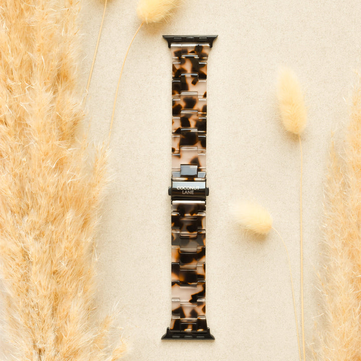 A Luxe Ivory Tort Apple Watch Strap from Coconut Lane is placed on a beige surface, surrounded by fluffy, beige ornamental grass on the left and right sides. The adjustable strap features silver connectors and prominently displays the brand name "Coconut Lane" on the clasp.