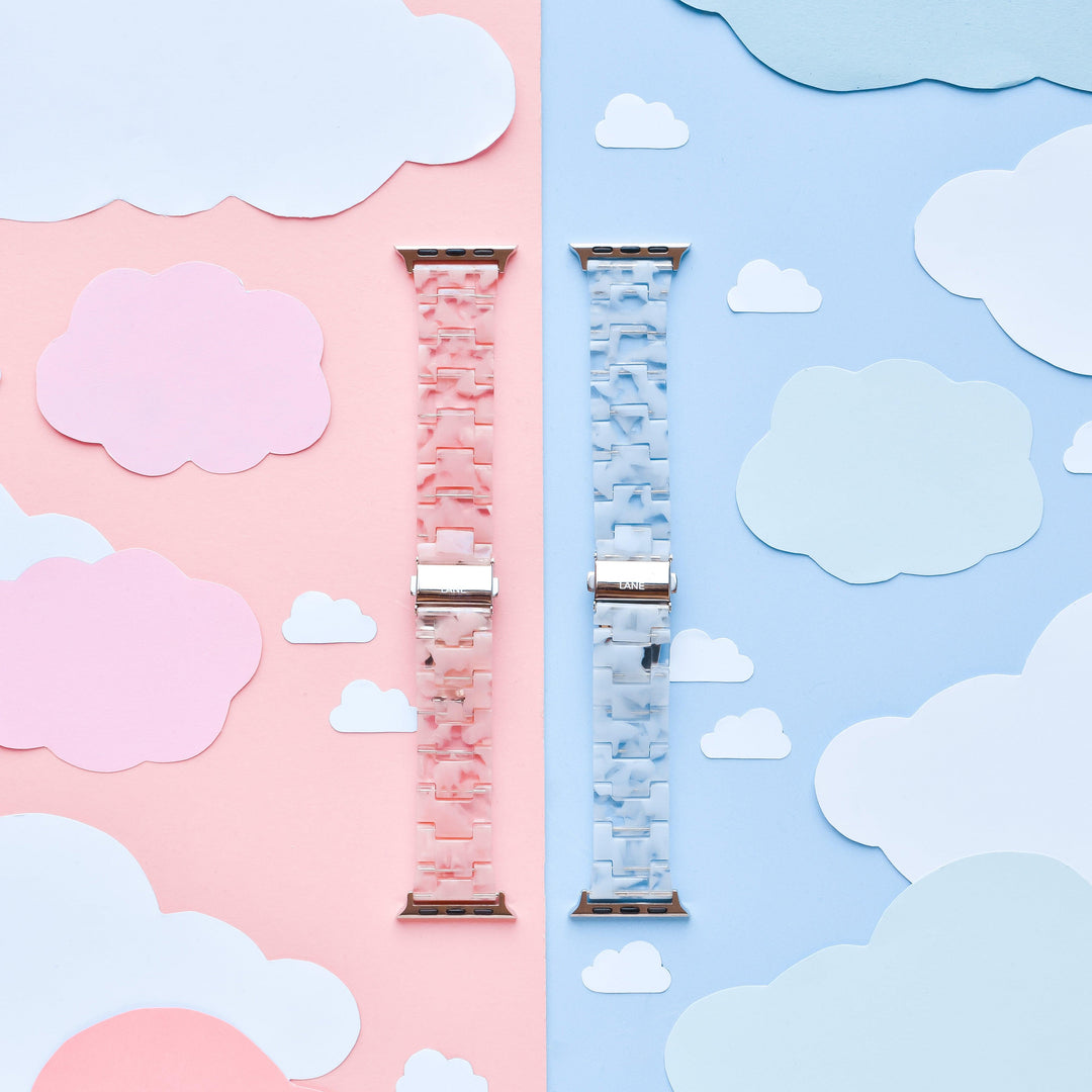 An image features two Apple Watch straps from Coconut Lane, including the Luxe Pink Clouds Apple Watch Strap and a blue counterpart, placed vertically side by side on a background divided into corresponding pink and blue sections. Each section is adorned with matching paper clouds, creating a girly and cute atmosphere. These durable resin bands add both style and function to your device.
