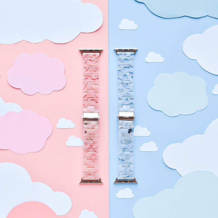 An image features two Apple Watch straps from Coconut Lane, including the Luxe Pink Clouds Apple Watch Strap and a blue counterpart, placed vertically side by side on a background divided into corresponding pink and blue sections. Each section is adorned with matching paper clouds, creating a girly and cute atmosphere. These durable resin bands add both style and function to your device.