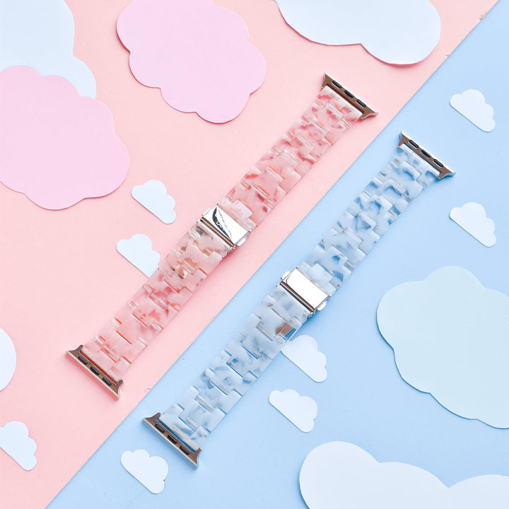 The image showcases two durable resin Apple Watch straps from Coconut Lane, featuring the Luxe Pink Clouds Apple Watch Strap in pink and another in blue. They are placed on a background divided into matching pink and blue sections, adorned with white and pink cloud cutouts. The bands have a marbled texture with metallic clasps, exuding a girly and cute aesthetic.