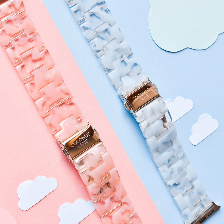 Displayed on a pastel pink and blue background are two Luxe Pink Clouds Apple Watch Straps by Coconut Lane, one in pink and the other in white. These girly and cute bands feature clasps engraved with "COCONUT LANE" and are set against a backdrop with small cloud cutouts.