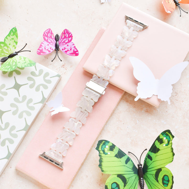 Introducing the Luxe Holo Butterfly Apple Watch Strap by Coconut Lane, showcased diagonally over pink and green tiles. This translucent, linked design is beautifully surrounded by decorative holographic butterflies in shades of green, pink, and white. The display exudes a light, airy, and creative aesthetic.