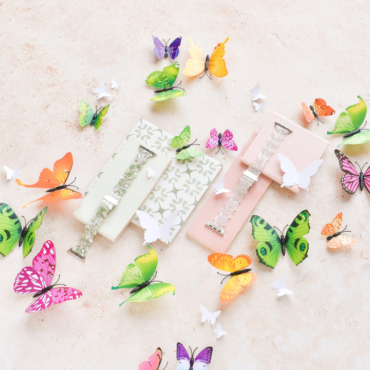 A pastel-themed image showcases vibrant holographic butterfly-shaped paper cutouts scattered around three Luxe Holo Butterfly Apple Watch Straps by Coconut Lane. The bands are elegantly displayed on decorative soft green, white, and pink packaging that complements the delicate aesthetic.