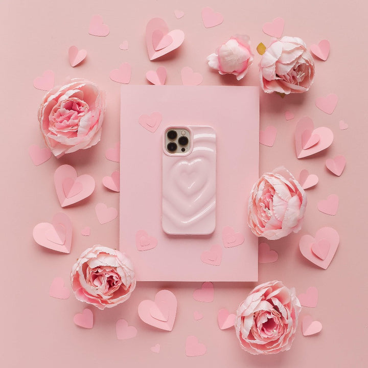 A pink smartphone adorned with Coconut Lane's Melting Heart Phone Case in Peony Pink is centered on a pink background. The phone is encircled by pink paper hearts of various sizes and clusters of light pink flowers, creating a romantic and delicate ambiance.