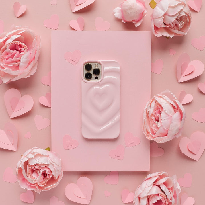 A Melting Heart Phone Case - Peony Pink by Coconut Lane lies on a larger pink rectangle, surrounded by pink paper hearts and pink peonies. The overall pink theme creates a soft, romantic aesthetic.