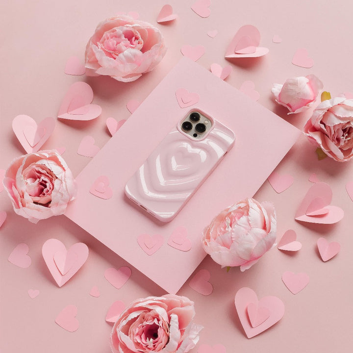 A Coconut Lane iPhone adorned with a Melting Heart Phone Case in Peony Pink rests on a soft pink rectangular sheet, surrounded by delicate pink peony flowers and heart-shaped paper cutouts. The backdrop is also a gentle shade of pink, creating a romantic and cohesive aesthetic.