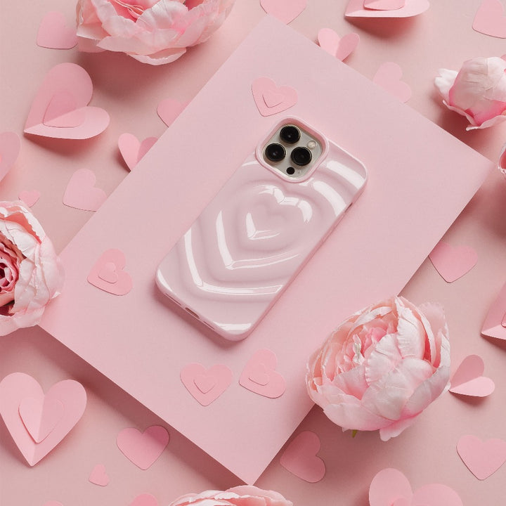 A smartphone with a Coconut Lane Melting Heart Phone Case in Peony Pink, featuring an adorable pink heart-shaped design, sits on a pink surface. It is surrounded by various sizes of pink paper hearts and delicate peony flowers. The overall theme is romantic, with a soft pink color scheme accentuated by the charming design.