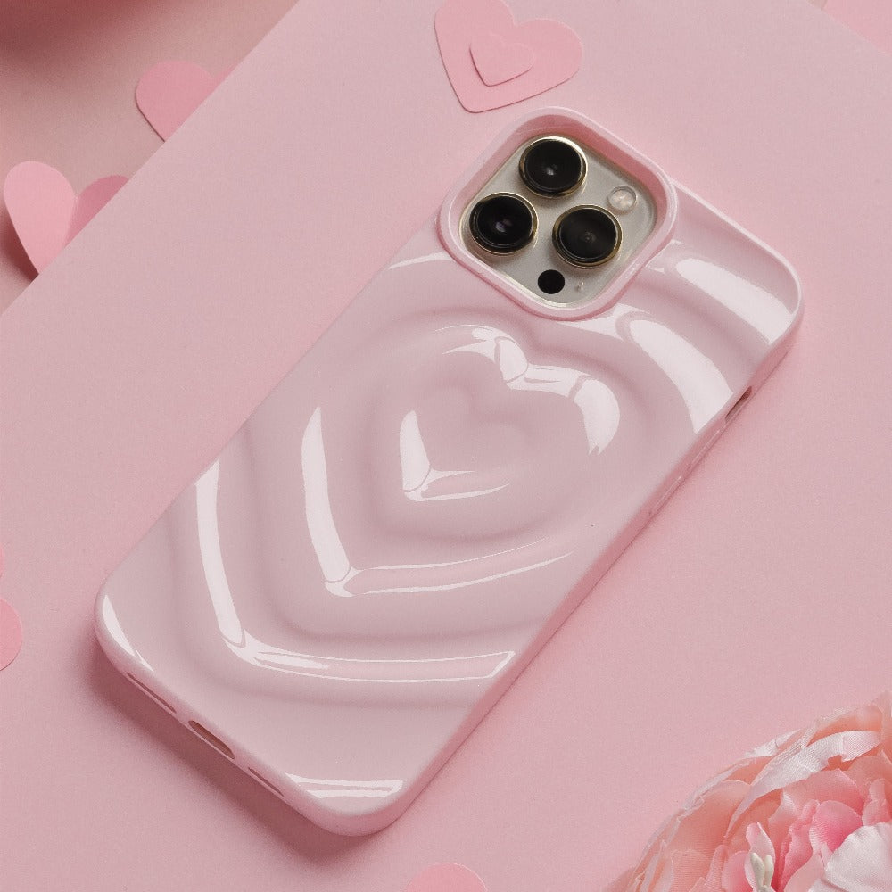 A Coconut Lane Melting Heart Phone Case in Peony Pink encases a smartphone resting on a pink surface. Multiple paper hearts are scattered around, enhancing the romantic and playful theme. The background features an array of pink shades and textures.