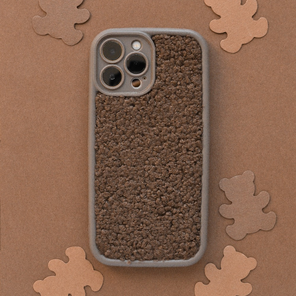 A Coconut Lane Cosy Teddy Phone Case in brown is placed flat on a brown surface. Surrounding the phone are several small, tan bear-shaped cutouts, creating a playful and cozy atmosphere while also adding an extra protective touch.