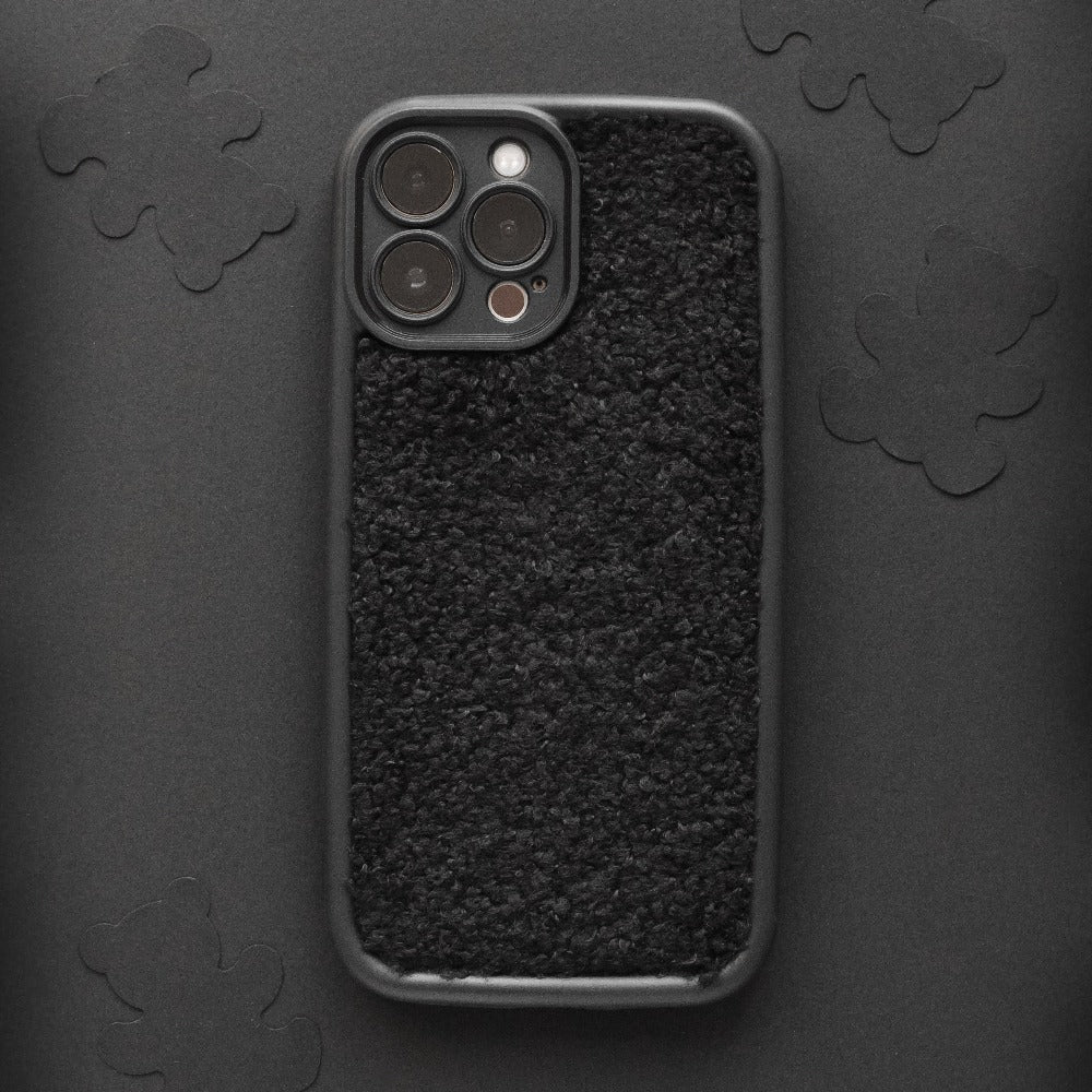 A smartphone with the Cosy Teddy Phone Case - Black from Coconut Lane is lying on a dark surface adorned with subtle floral patterns. The protective phone case features a fuzzy, fleecy texture, and the phone's camera lenses are visible in the upper left corner.