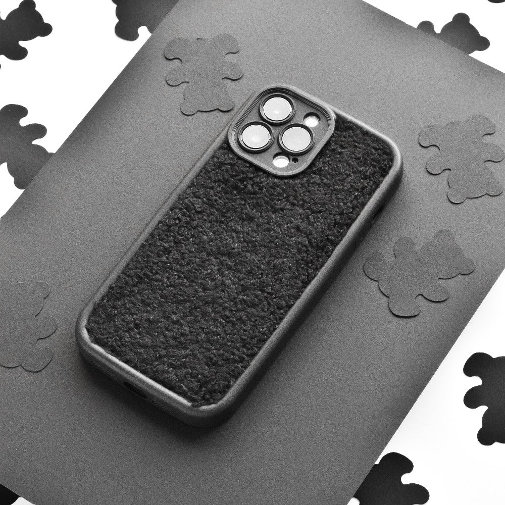 A black Cosy Teddy Phone Case from Coconut Lane is displayed on a grey surface. The protective phone case has cut-outs for the camera and buttons. The background features scattered black cloud-shaped cut-outs on white paper, creating a contrasting pattern.