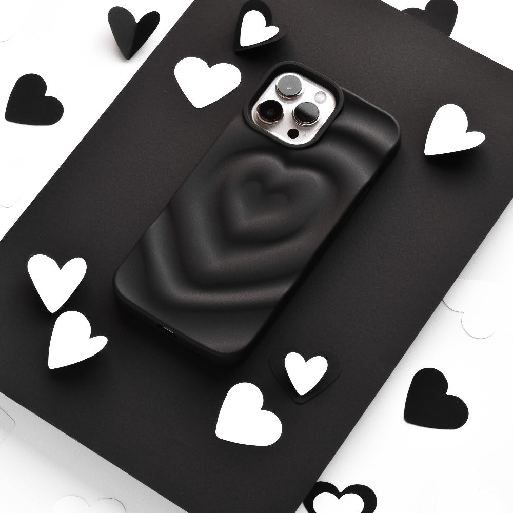 A smartphone encased in Coconut Lane's Melting Heart Phone Case - Matte Black sits on a black surface, surrounded by scattered black and white heart-shaped cutouts. The image emphasizes a monochromatic theme with a focus on the phone and the heart shapes.