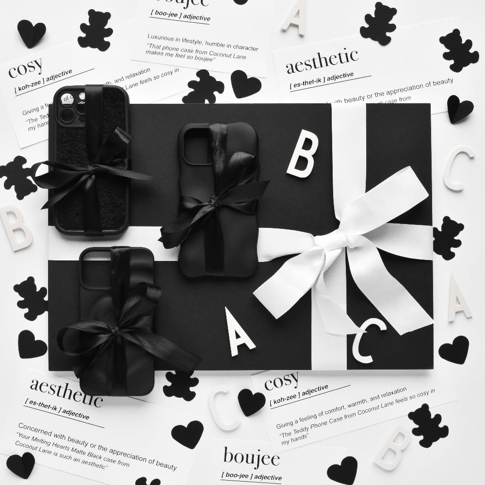 A black-and-white flat lay image features phone cases adorned with bows, decorative letters, hearts, teddy bear cutouts, and cards showcasing words and their definitions ("cosy," "aesthetic," "boujee"). The monochromatic theme is highlighted by the textured Cosy Teddy Phone Case in Black from Coconut Lane, enhancing the contrasting black-and-white elements.
