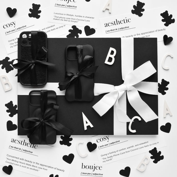 A black-and-white flat lay image features phone cases adorned with bows, decorative letters, hearts, teddy bear cutouts, and cards showcasing words and their definitions ("cosy," "aesthetic," "boujee"). The monochromatic theme is highlighted by the textured Cosy Teddy Phone Case in Black from Coconut Lane, enhancing the contrasting black-and-white elements.