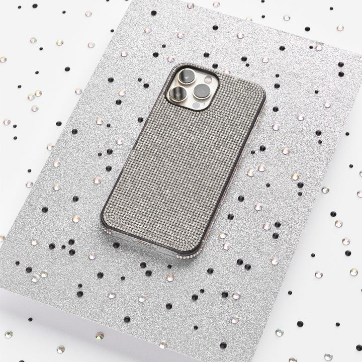 A silver iPhone adorned with the Coconut Lane Disco Sparkle Phone Case - Silver, showcasing a grid pattern, rests on a glittery silver surface. The background is sprinkled with small black and silver sequins, setting a festive and glamorous scene.