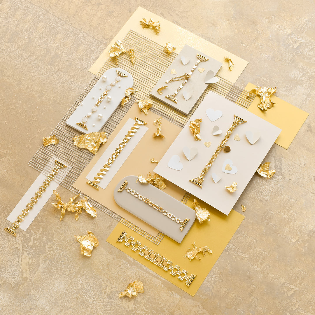 A flat lay of various gold and white accessories on a beige surface. The accessories include Coconut Lane's Simple Gold Bracelet Apple Watch Strap, gold chains, and white elements such as hearts and geometric shapes. Gold foil pieces are scattered around, adding a luxurious touch to the composition.
