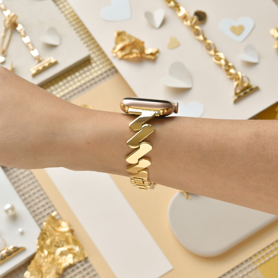 A close-up of a wrist wearing an Apple Watch fitted with the Get Wavy Apple Watch Strap - Gold by Coconut Lane, showcasing its bracelet-style zigzag link design. The background features scattered gold and white decorative elements, including hearts, pearls, and gilded fragments, creating a luxurious and elegant setting.