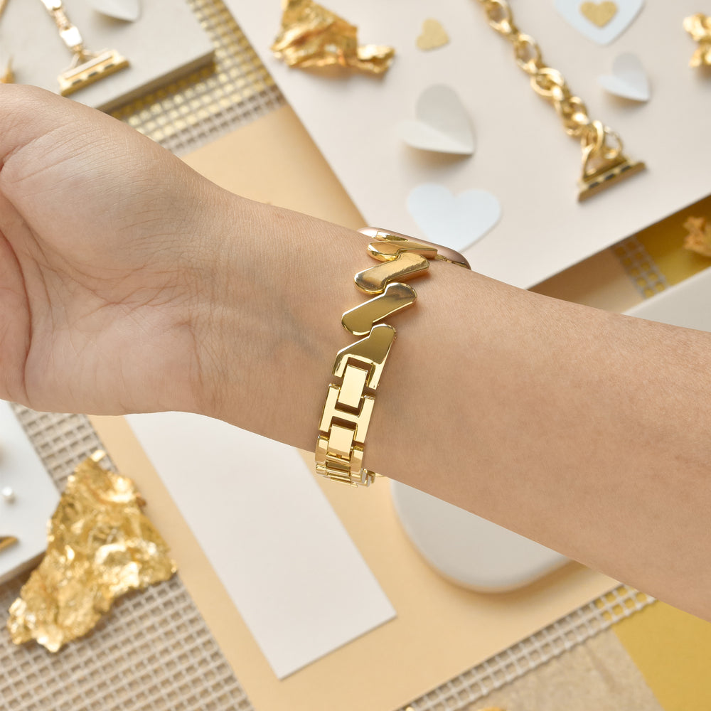A wrist adorned with a gold bracelet featuring interlocking heart and rectangular links, complemented by the Get Wavy Apple Watch Strap - Gold from Coconut Lane. The background has materials used for jewelry making, such as gold foil and paper hearts, scattered on a beige and white surface.