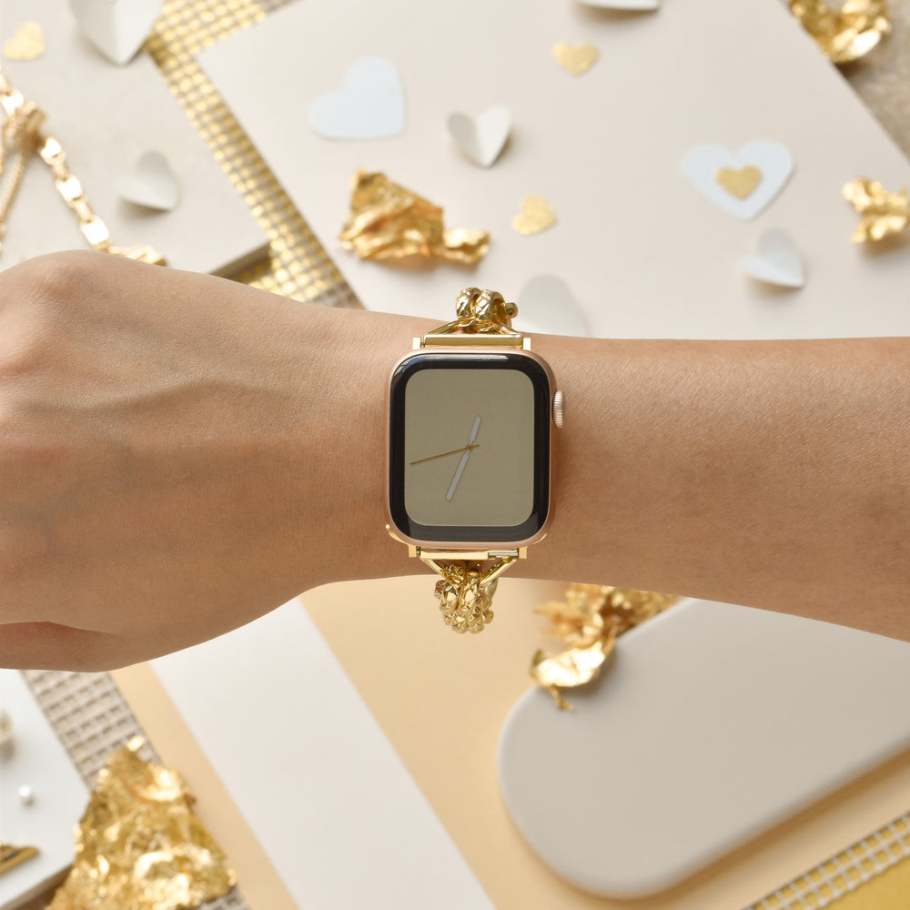 A close-up of a person's wrist wearing the Coconut Lane Chain Bracelet Apple Watch Strap - Gold, featuring an adjustable stainless steel metal design. The background showcases beige and gold decorative elements, including hearts and crumpled gold foil pieces. The time on the watch face shows approximately 7:00.