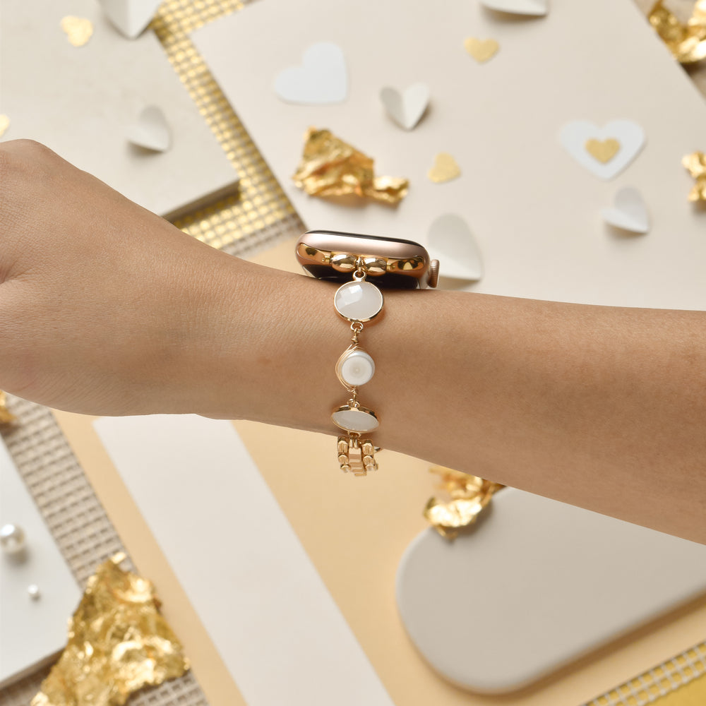 A person’s wrist adorned with the Cute Stone Bracelet Apple Watch Strap by Coconut Lane, featuring delicate gold chains and oval-shaped white gemstones. The background includes scattered white heart shapes, gold foil accents, and neutral-toned papers, creating a sophisticated and artistic presentation of this stylish watch strap.