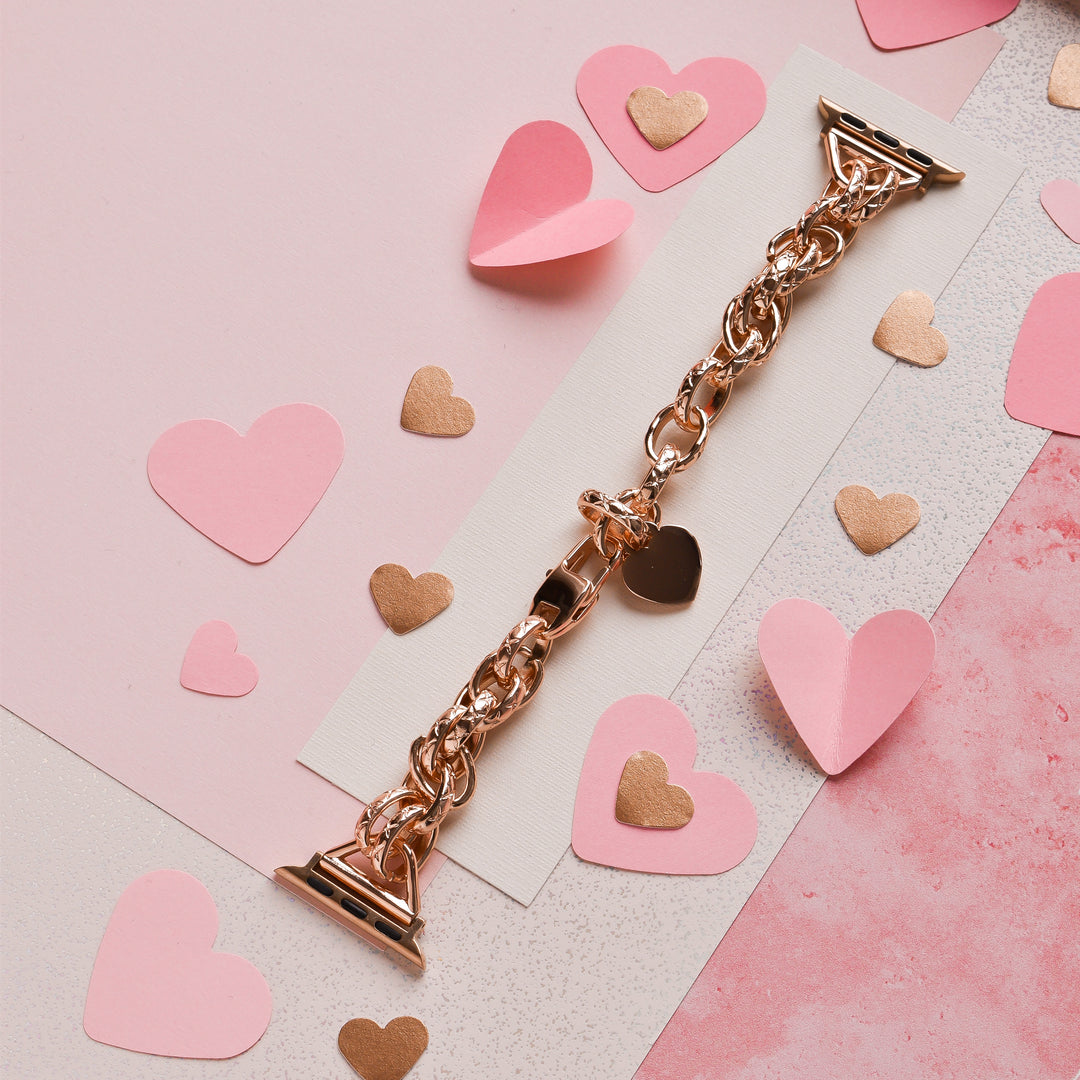 A rose gold Chain Bracelet Apple Watch Strap from Coconut Lane is placed on a pink and white background. Surrounding the watch strap are various cut-out paper hearts in pink and gold, creating a romantic and festive setting.