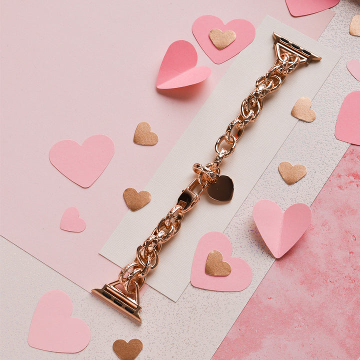 A rose gold Chain Bracelet Apple Watch Strap from Coconut Lane is placed on a pink and white background. Surrounding the watch strap are various cut-out paper hearts in pink and gold, creating a romantic and festive setting.