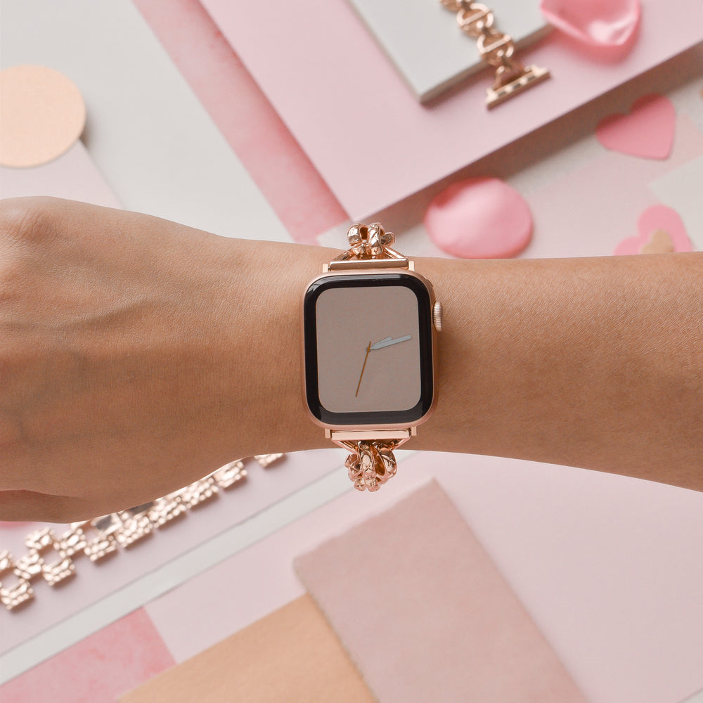 A person's wrist is adorned with a Coconut Lane Chain Bracelet Apple Watch Strap in rose gold, complementing the stylish smartwatch. The watch face displays a minimalistic analog clock design, set against a backdrop of pink and neutral-colored papers and decorative elements that create a visually appealing scene.