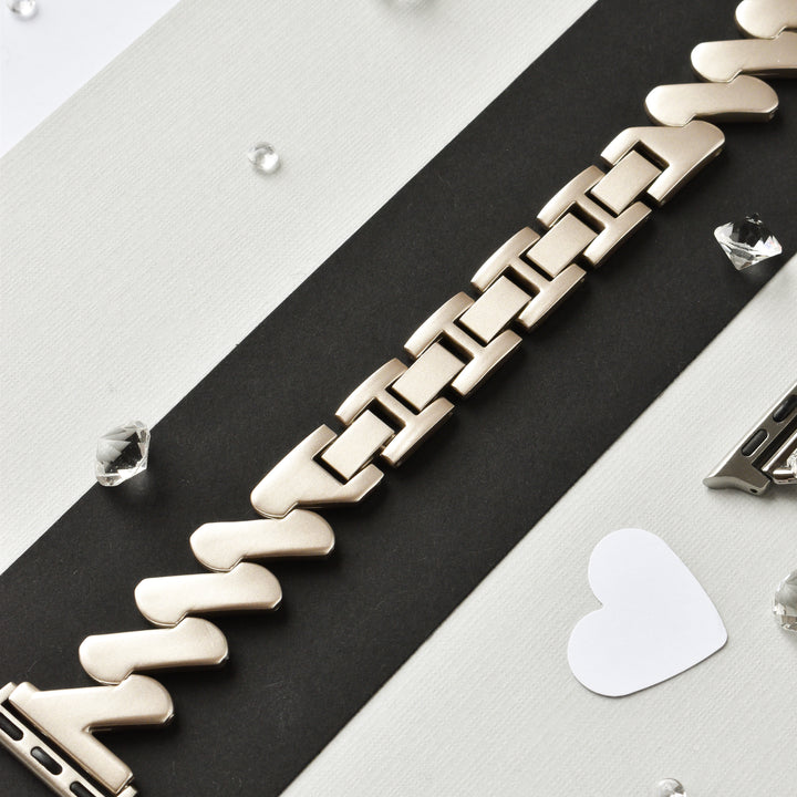 A close-up shot of the Get Wavy Apple Watch Strap - Starlight by Coconut Lane, laid diagonally on a black and white striped surface, surrounded by small, clear gemstones and a white heart cutout. The stainless steel adjustable watch band's interlocking links feature a matte finish. This strap is compatible with ALL Apple Watch models.