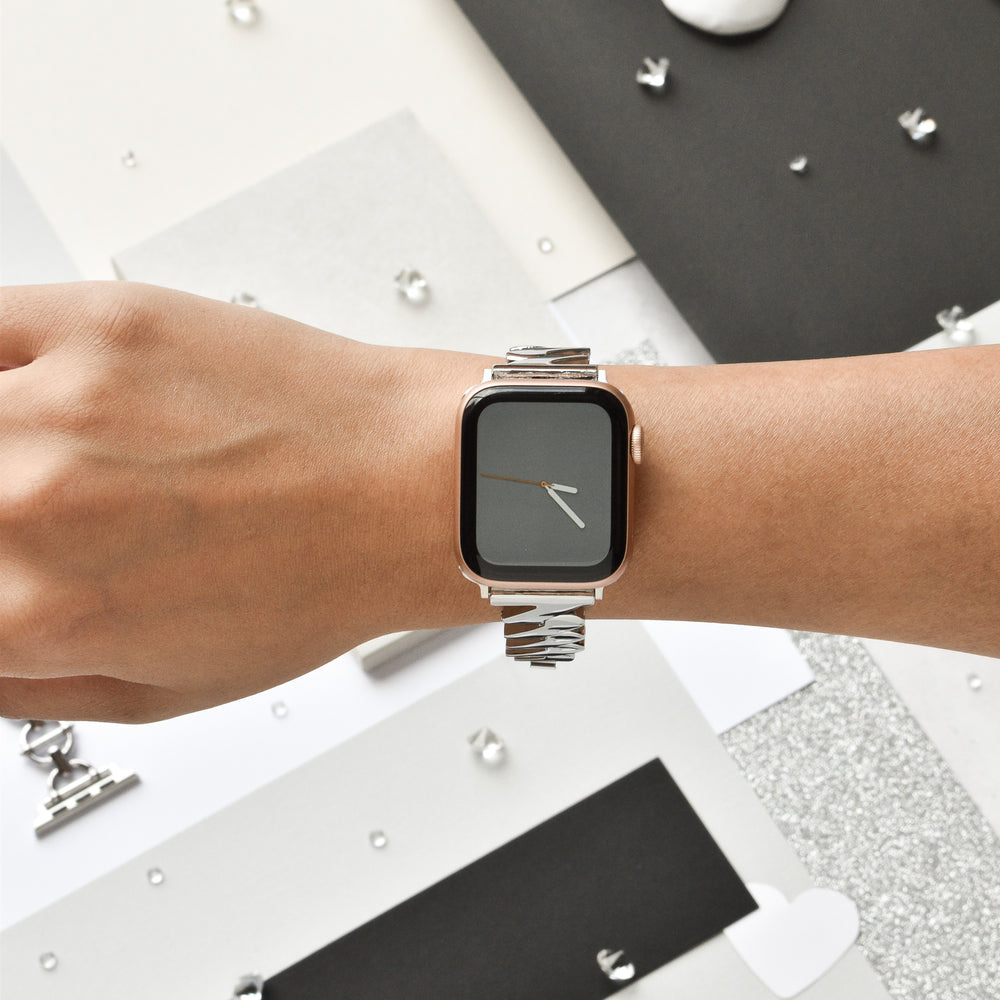 Close-up of a person's wrist wearing a silver smart watch with a rectangular face and minimalist design, featuring a black screen displaying a simple clock with white hands. The Get Wavy Apple Watch Strap - Silver by Coconut Lane is made of sleek stainless steel. The background consists of geometric patterns and abstract shapes in monochrome tones.