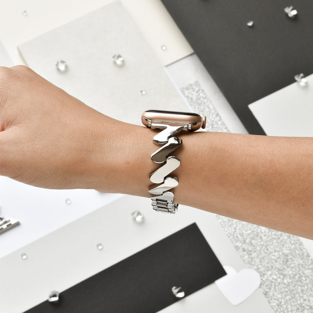 A person's wrist adorned with the Get Wavy Apple Watch Strap in Silver by Coconut Lane, featuring a sleek and abstract wavy bracelet design in stainless steel. The background showcases various color blocks and textured papers in shades of black, white, and gray, along with scattered decorative rhinestones.