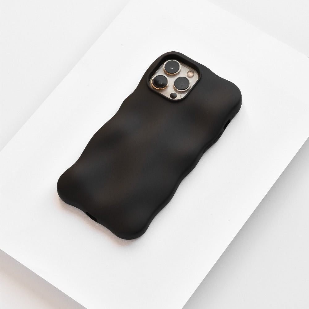 A black iPhone with a Coconut Lane 3D Boujee Phone Case in Matte Black rests on a white surface. The phone's camera array is visible on the back, featuring three lenses and a flash. The matte black finish of the extra protective, minimalist design emphasizes the soft, wavy texture and shape of the case.