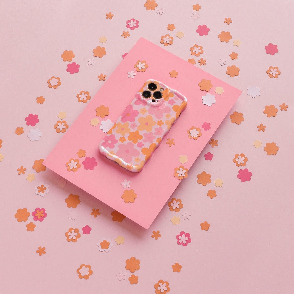 Coconut Lane's Wavy Phone Case - Pink & Orange Bloom, adorned with an extra protective pink and orange floral design, lies on a slightly darker pink sheet surrounded by small, scattered paper flowers in shades of pink, peach, and orange on a light pink background.