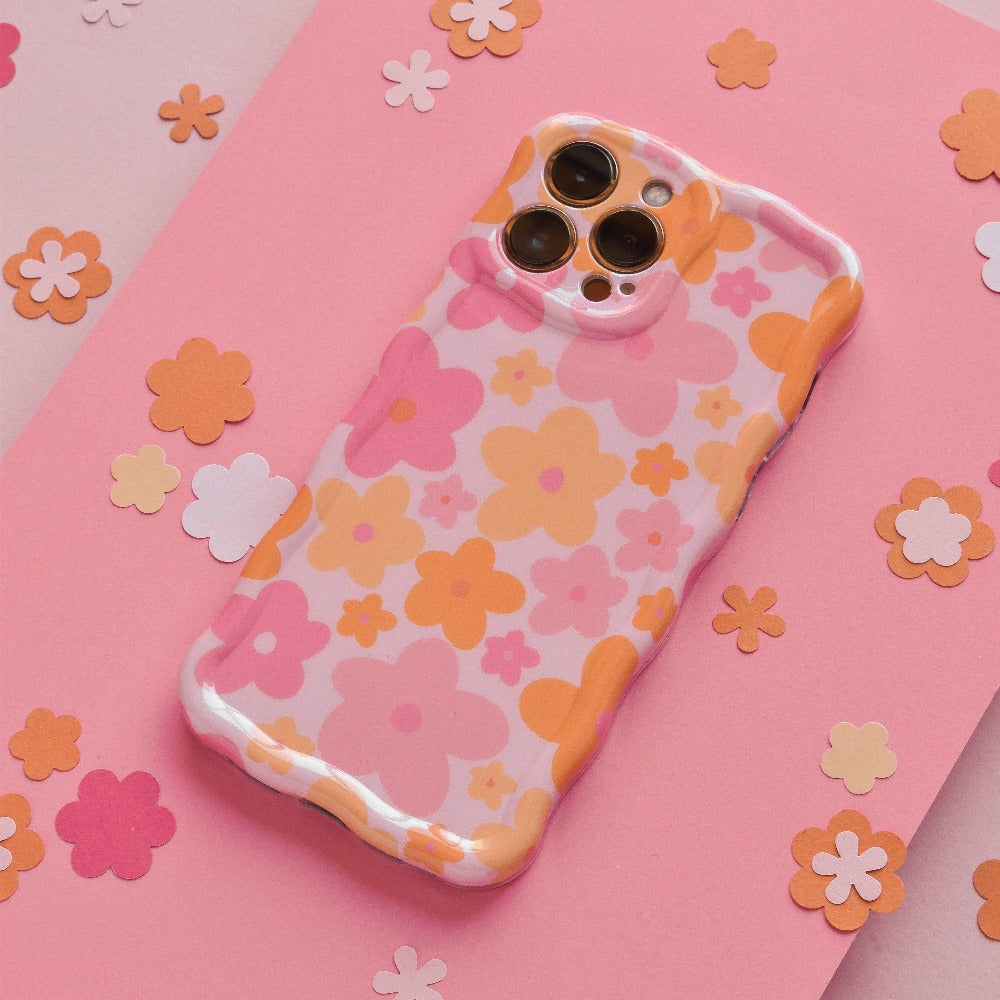 A Coconut Lane Wavy Phone Case - Pink & Orange Bloom lies on a pink surface scattered with small, flower-shaped cutouts in various shades of pink and orange. The case's extra protective design complements the cohesive, vibrant, and playful floral aesthetic.
