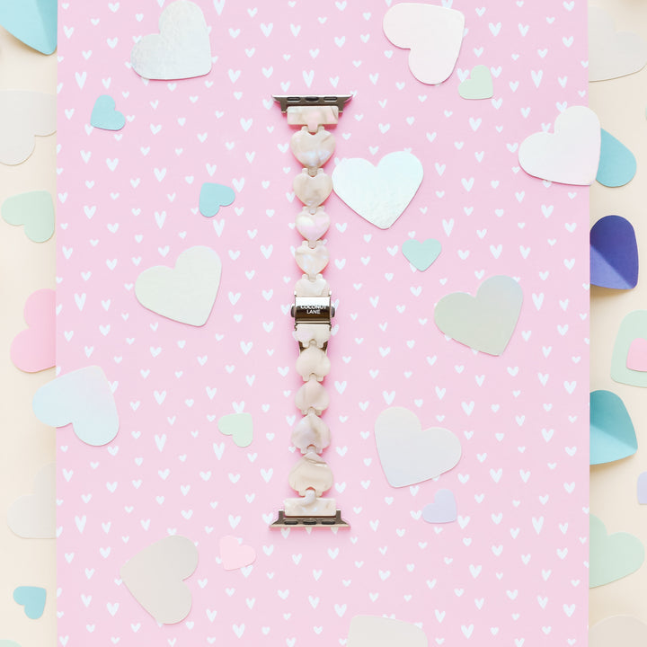 A pearly-beaded charm bracelet with heart-shaped beads lies vertically on a pink background adorned with white heart patterns. Scattered larger pastel-colored hearts frame the edges, creating a whimsical and romantic setting. This delicate design perfectly complements the Luxe Hearts Apple Watch Strap - Rose Quartz from Coconut Lane.