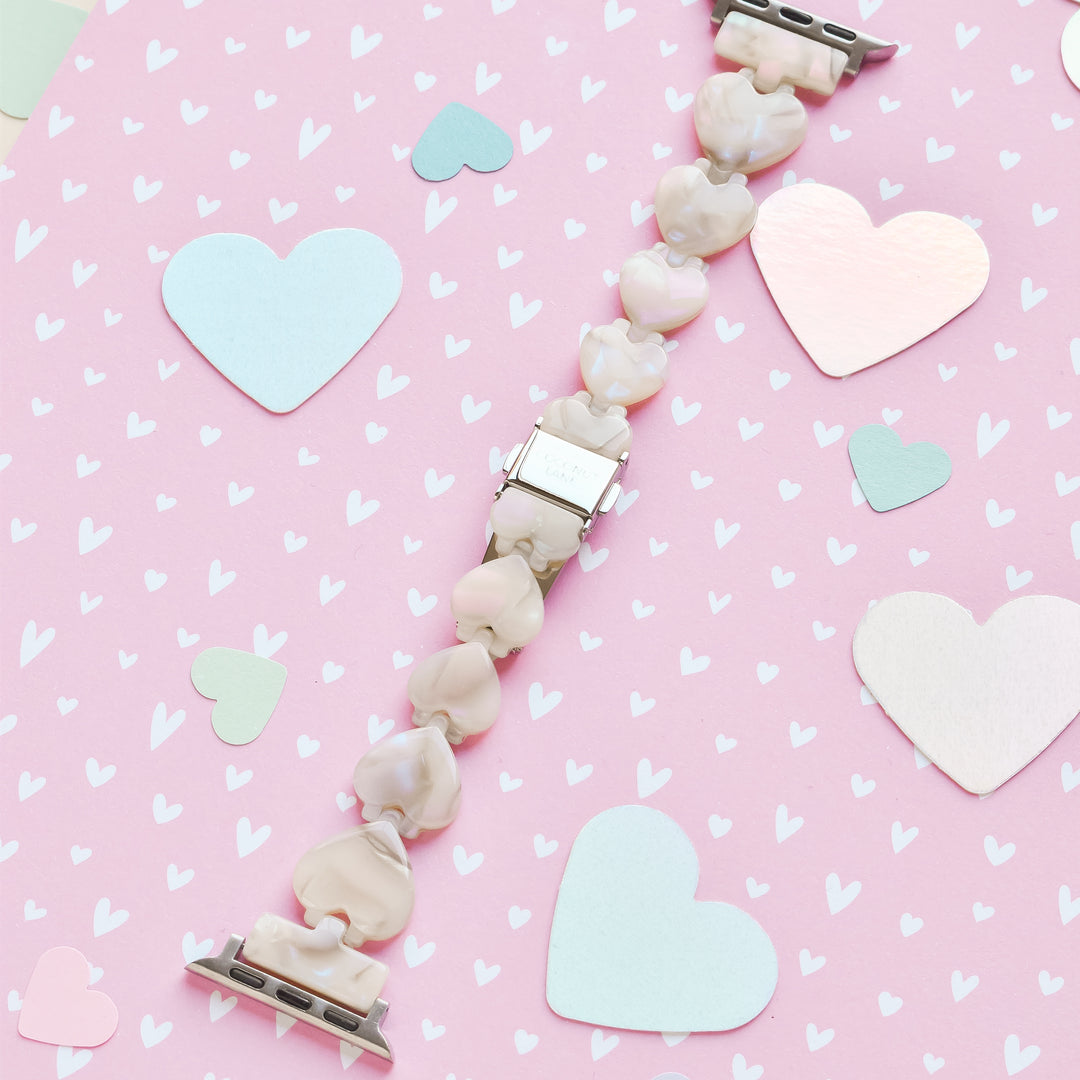 A bracelet featuring light pink heart-shaped beads and silver clasps was laid out on a pink background adorned with hearts of various sizes and colors, including shades of blue, white, metallic pink. The display is harmoniously paired with the Luxe Hearts Apple Watch Strap - Rose Quartz by Coconut Lane for an elegant touch.