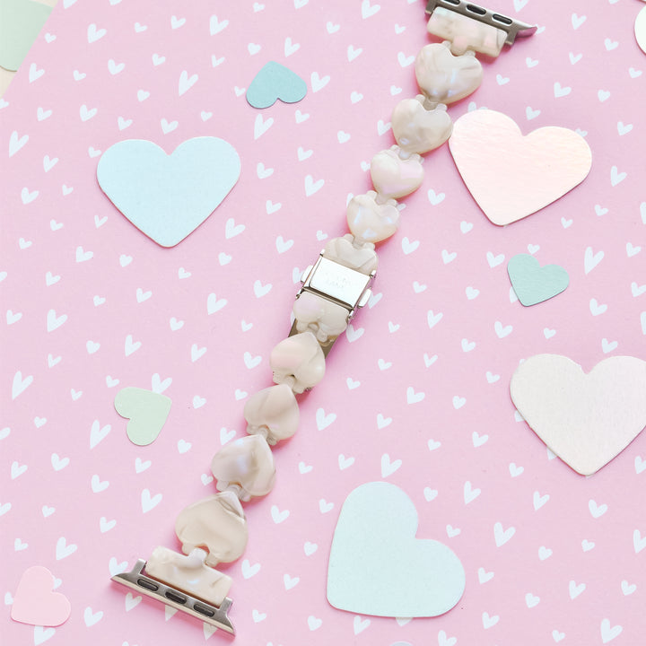 A bracelet featuring light pink heart-shaped beads and silver clasps was laid out on a pink background adorned with hearts of various sizes and colors, including shades of blue, white, metallic pink. The display is harmoniously paired with the Luxe Hearts Apple Watch Strap - Rose Quartz by Coconut Lane for an elegant touch.