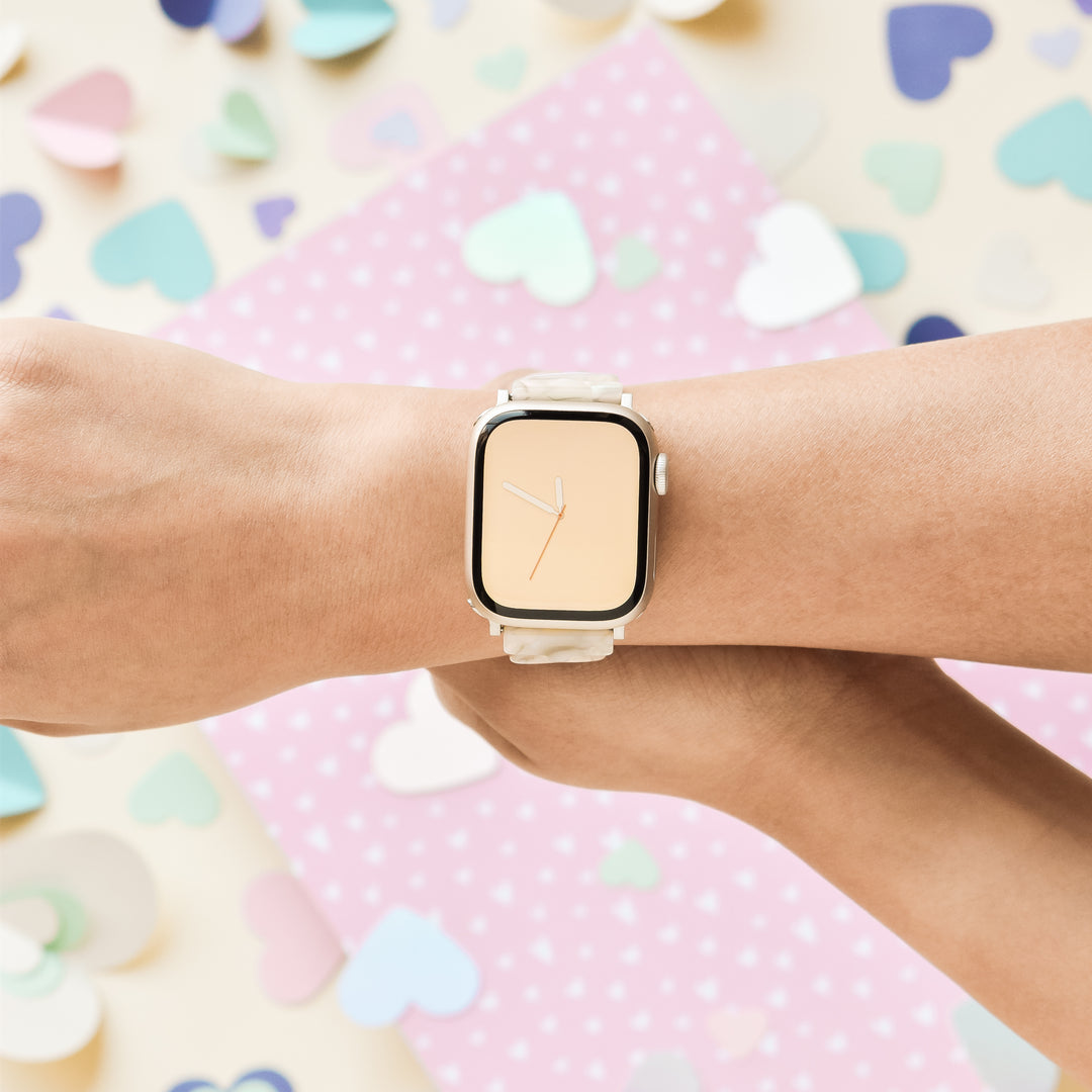 A person wearing a smartwatch with a pastel yellow screen and the Luxe Hearts Apple Watch Strap - Rose Quartz from Coconut Lane on their wrist. The background is dotted with colorful, heart-shaped paper cutouts and a pink polka-dotted sheet, creating a cheerful and whimsical atmosphere.