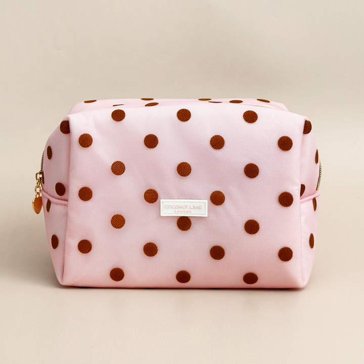 Pink mesh makeup bag with brown polka dots, a gold top zip with a heart-shaped zip pull, and a white Coconut Lane London logo badge on the front, shown on a beige background.