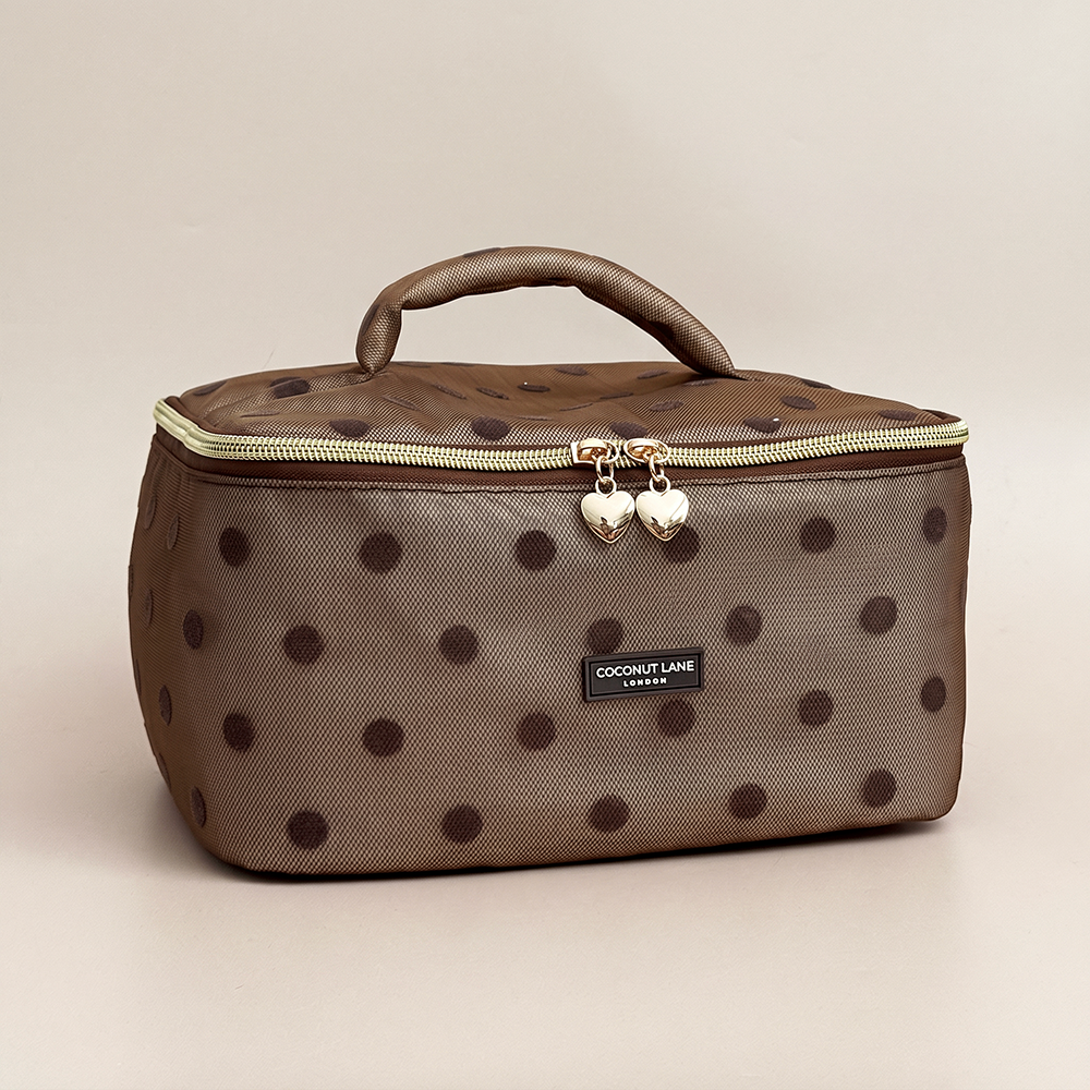 Brown mesh vanity case with dark brown polka dots, a padded top handle, gold zip with two heart-shaped zip pulls, and a black Coconut Lane London logo badge on the front, shown on a beige background.