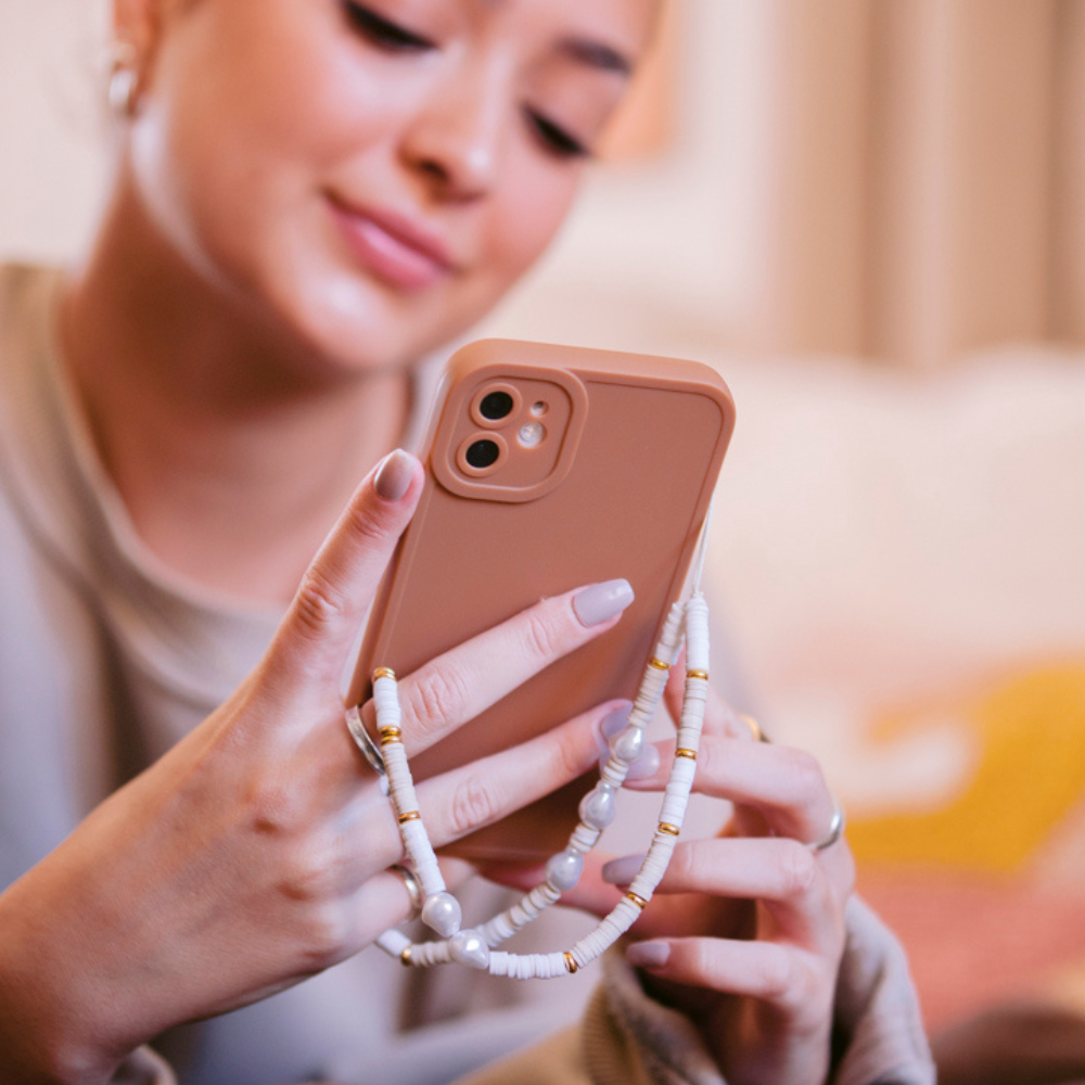 A person holds a smartphone adorned with Coconut Lane's Beaded Phone Strap in Nude & Pearl and a beige phone case. They are smiling while sitting indoors, their nails manicured in a light color. The background is softly blurred, contributing to the warm and cozy atmosphere.