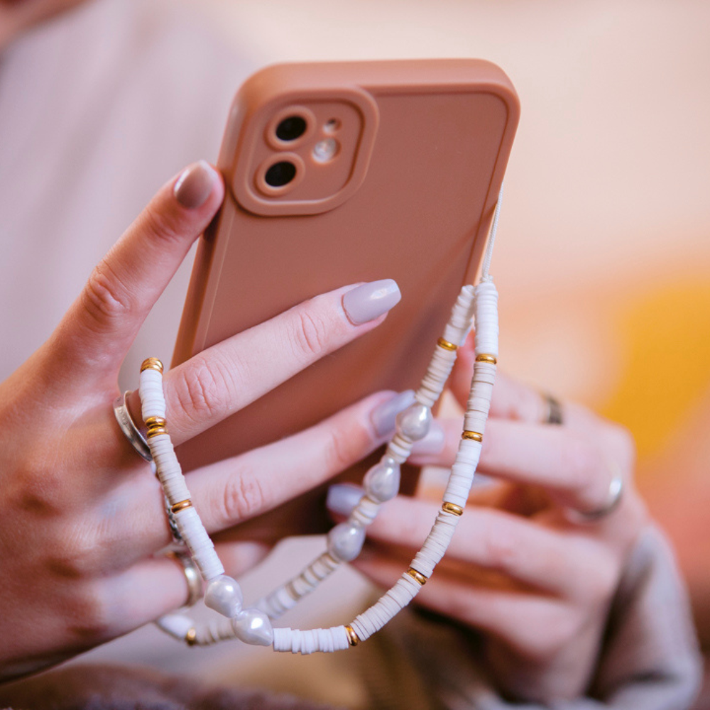 A close-up of hands holding a peach-colored phone case, adorned with Coconut Lane’s Beaded Phone Strap - Nude & Pearl, featuring white and gold beads. The person has light purple nail polish and is wearing silver rings. This stylish accessory adds an extra touch of charm to the overall look.