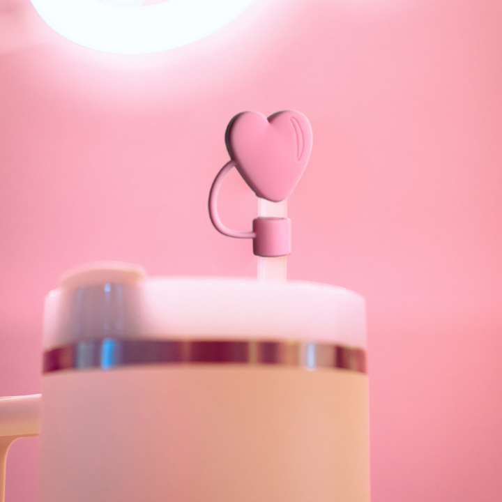 A close-up view of Coconut Lane's tumbler, featuring a pink heart-shaped lid decoration from their Pink Heart Straw Cover collection. The background is a soft pink, creating a cohesive and aesthetically pleasing effect. The flexible design includes a silicone straw cover for added convenience.