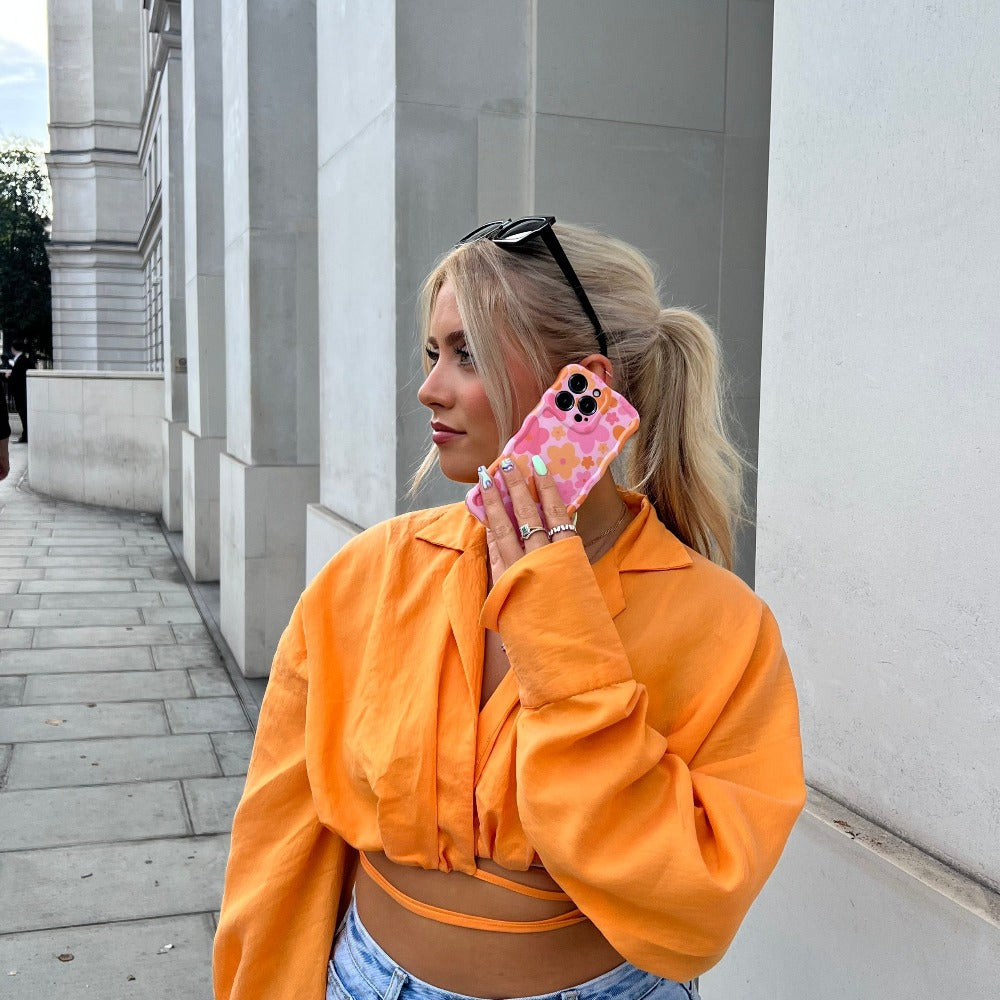 A person with blonde hair, wearing black sunglasses on their head, an orange button-up shirt, and blue jeans, is holding a smartphone with a Coconut Lane Wavy Phone Case - Pink & Orange Bloom to their ear. They are standing on a street with light-colored buildings in the background.